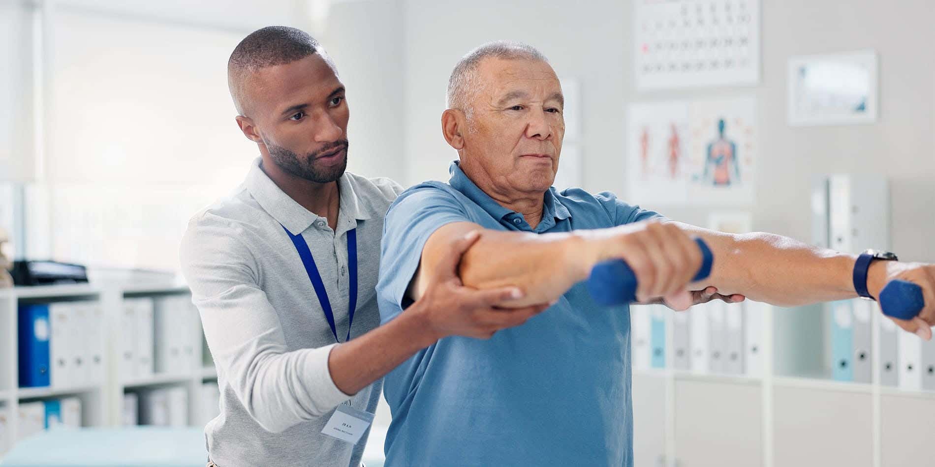A physical therapist supports an older man’s arm as he performs a shoulder exercise with small dumbbells in a clinic.