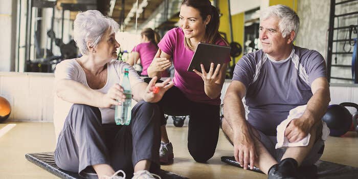 A fitness professional kneels between two older adults seated on exercise mats, using a tablet as they talk in a gym.
